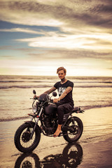 Obraz na płótnie Canvas handsome tattooed biker riding motorbike on ocean beach and looking at camera