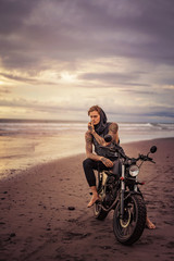 Obraz na płótnie Canvas handsome tattooed biker posing on motorcycle on ocean beach