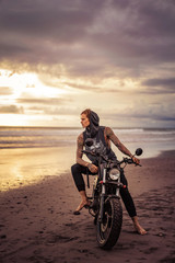 Fototapeta na wymiar handsome tattooed man sitting on motorcycle on ocean beach and looking away