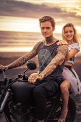 young heterosexual couple sitting on motorcycle and looking at camera at beach