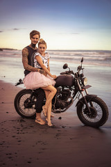 couple hugging on motorcycle on ocean beach during beautiful sunrise and looking at camera