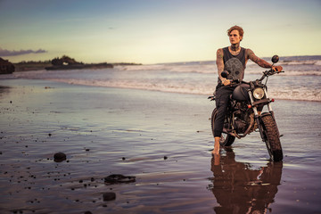 tattooed man sitting on motorbike on ocean beach and looking away