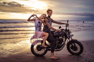 Fototapeta na wymiar girlfriend leaning on boyfriend on motorcycle and showing heart with fingers on ocean beach
