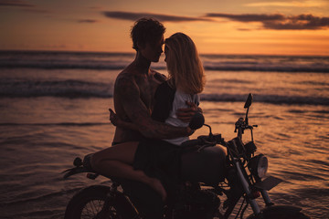 Obraz na płótnie Canvas passionate boyfriend and girlfriend hugging on motorbike at beach during sunset