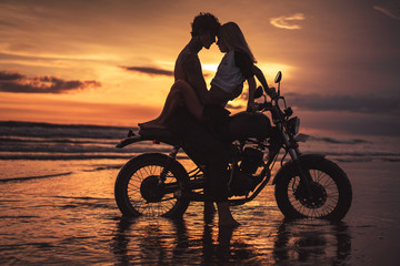 Obraz na płótnie Canvas passionate couple hugging and touching with foreheads on motorbike at beach during sunset