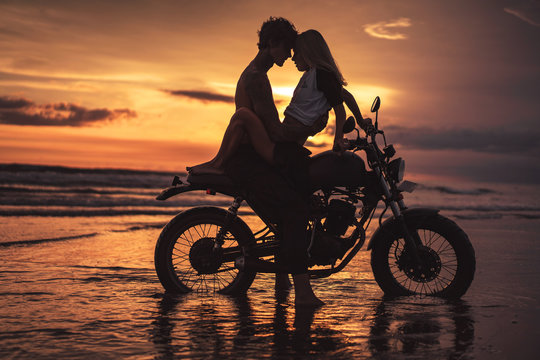 passionate couple hugging and touching with foreheads on motorbike at beach during sunset