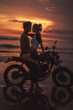 passionate couple hugging and going to kiss on motorcycle at beach during sunset