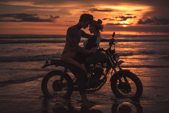 passionate couple hugging and touching with foreheads on motorcycle at beach during sunset