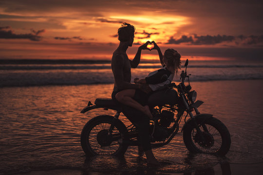 side view of couple making heart with fingers on motorcycle at beach during sunset