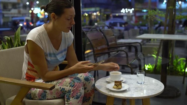 A woman in a cafe with a smartphone drinks coffee and eats a tiramisu cake