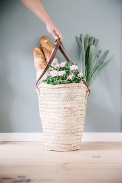 A woman hand holding a rush or wicker basket with light pink roses, with two baguettes and a couple of leeks.