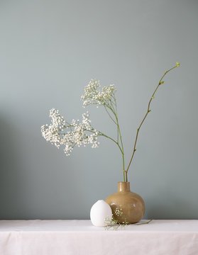 A floral setting with ceramic vases filled with baby's breath and a linden branch on a white table top against a muted grey green wall.