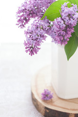 Close-up part of white ceramic pot with purple blooming lilac standing on wooden round board, on table.