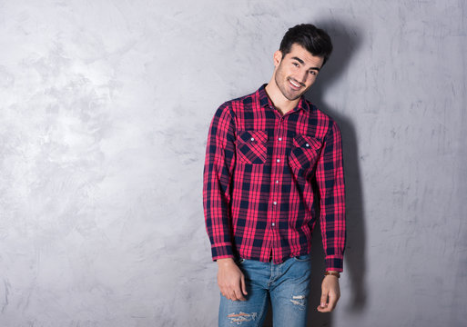 A happy handsome young man in a red checkered shirt standing in front of a grey wall in a studio.