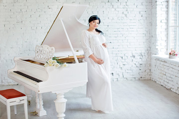 Happy pregnant girl in white dress stands near white piano