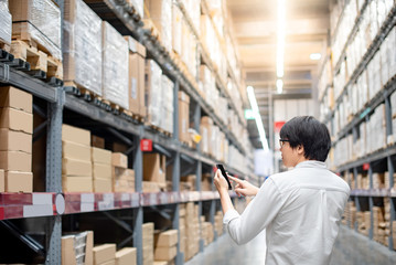 Young Asian man checking the shopping list on his smartphone between product shelves. warehouse shopping lifestyle concept - obrazy, fototapety, plakaty