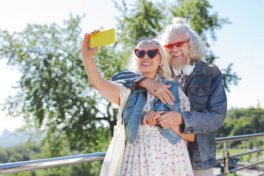Happy moments. Joyful aged woman standing with her husband while taking selfies with a smartphone