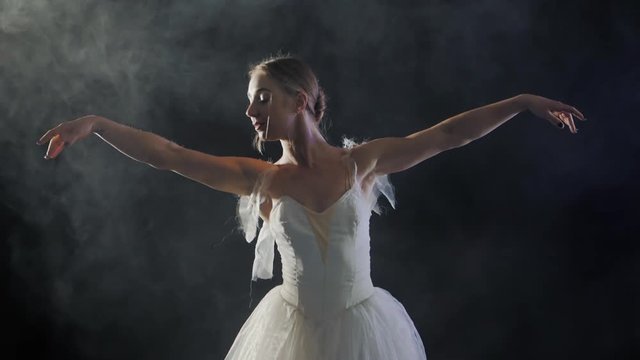 Ballerina is practicing her moves in dark studio. Young girl dancing with air white dress tutu and smiling. Gracefulness and tenderness in every movement. Slow motion.