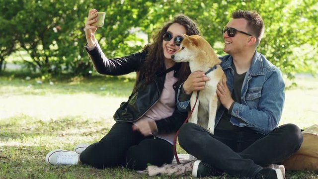 Happy couple is taking selfie with their dog using smartphone sitting on grass in the park, people are posing, talking and petting shiba inu puppy.