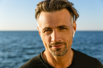 Fototapeta premium close-up portrait of smiling adult man with wireless earphones on seashore looking at camera