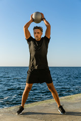 handsome adult sportsman working out with fit ball and holding it above head on seashore