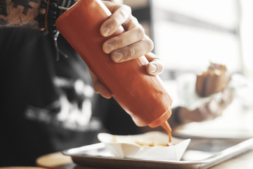 Young bearded hipster man pours ketchup on french fries close up.