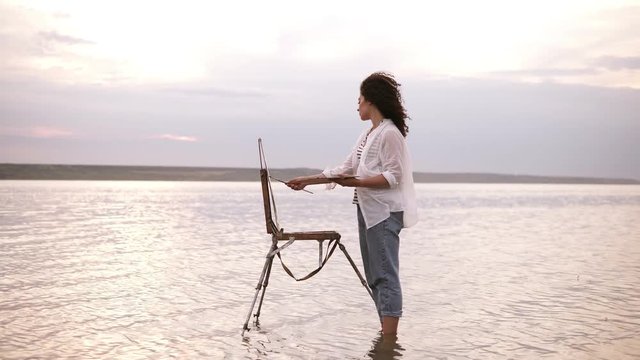 Gorgeous view of a young curly brunette standing in the water with easel and draing her the surrounding landscape. Wearing a blue jeans and white shirt. Full length