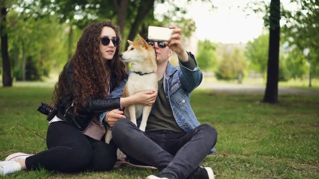 Handsome young man is taking selfie with his pretty wife and cute dog, all wearing sunglasses. Guy is holding smartphone taking pictures and posing.