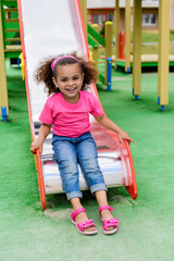  smiling curly african american little kid sliding down from hill at playground