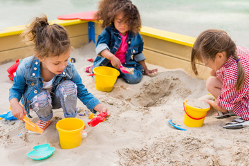 three multiethnic little children playing with plastic scoops and buckets in sandbox at playground - obrazy, fototapety, plakaty
