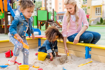 selective focus of mother playing with daughter and her little friend in sandbox at playground