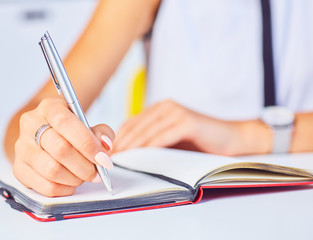 Young woman working at her desk taking notes closeup. Focus on hand writing on a notepad.