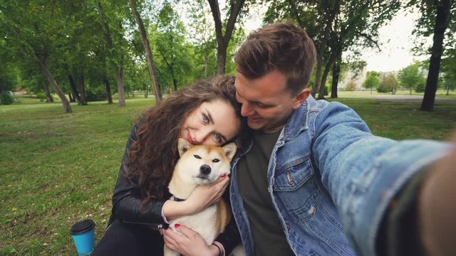 Young wife and husband are taking selfie with adorable dog kissing and hugging each other and the animal. Point of view shot of happy people, pet and green park.