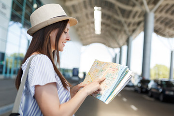 Young smiling traveler tourist woman in hat with backpack searching route in paper map at international airport. Female passenger traveling abroad to travel on weekends getaway. Air flight concept.
