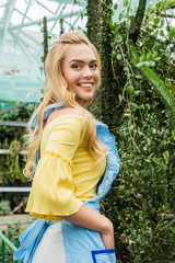 side view of beautiful girl in apron smiling at camera while standing near green plants