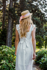 back view of attractive girl in white dress and wicker hat walking in forest