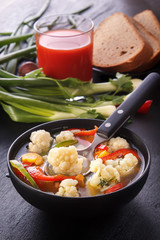 Vegetable soup of cauliflower, carrots, tomato, pepper in a black plate with a spoon on a black background with greens, rye bread and a glass of tomato juice
