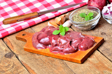 Raw chicken liver on a wooden cutting board, fresh garlic, dried marjoram, oregano, onion, knife and red checkered tablecloth in the background