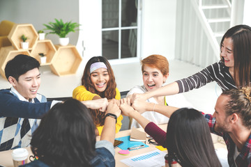 Fototapeta premium Multiethnic young team stack hands together as unity and teamwork in modern office. Diverse group togetherness collaboration or friends huddle concept. Startup business team building with soft tone.