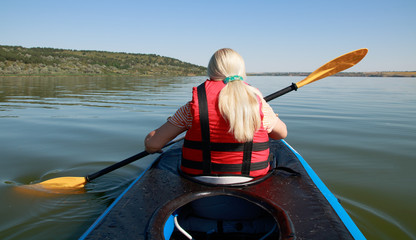 blond female kayaking on the lake, view from the back. green water, banks. unrecognizable people