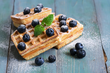 Homemade Belgian waffles with blueberries are sprinkled with powdered sugar on a green wooden rustic background. Selective focus.