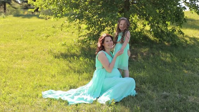 Happy pregnant mother with a little daughter in the same dress sitting on the grass in the Park, they laugh.