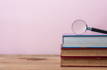 magnifying glass and pile of books on wooden table, pink pastel background - Powered by Adobe