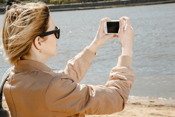 The girl picks up the city over the river on the phone. Young woman with a mobile phone near the river. Panorama of the city. Mobile photography. stylish woman with shoulder bag and sunglasses on the