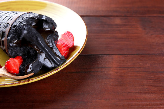 Black ice cream and sliced strawberries on a yellow plate, ice cream with chocolate in waffle cone with a wooden spoon, summer dessert with red berries on a wooden background, cold milk product - Powered by Adobe