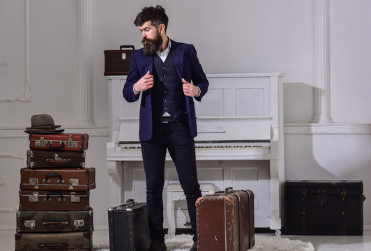 Baggage and travelling concept. Macho stylish on thoughtful face standing near pile of vintage suitcase. Man, traveller with beard and mustache with luggage, luxury white interior background.