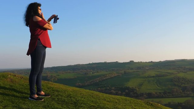 Long haired woman on the top of the hill lit by evening sun is taking landscape pictures at sunset 