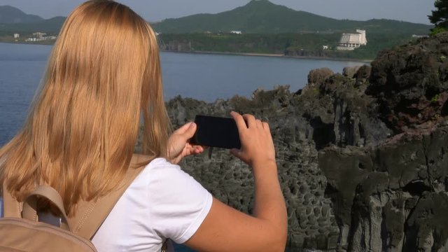 Caucasian Female Tourist Taking Photo of Daepo Jusangjeolli Cliff on Jeju Island (South Korea) with Mobile Phone. Active Tourism in Asia
