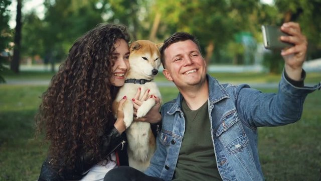 Slow motion of handsome guy taking selfie with his wife and dog in the park, cheerful man is posing while pretty woman with curly hair is hugging shiba inu puppy.