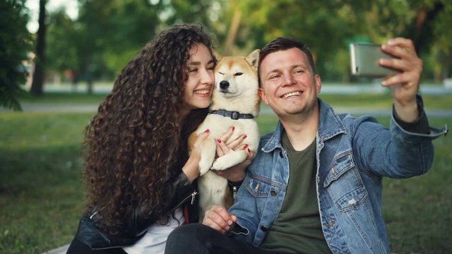 Slow motion of happy pair girl and guy taking selfie with cute dog posing and kissing animal holding smartphone. Modern technology, parks and leisure concept.